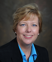 Patricia Crawels Patricia Crawels headshot, Caucasian business woman wearing a black suit with a light blue button down shirt pictured in front of a beige backdrop
