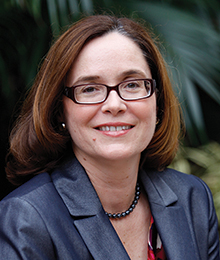 Debra Henley Debra Henley headshot, Caucasian woman photographed outdoors in front of a plant, she's wearing glasses and a navy suit
