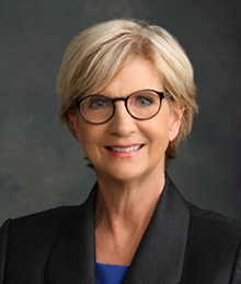 Elizabeth King Elizabeth King headshot, Caucasian middle aged woman wearing glasses and a black suit, pictured in front of a solid gray background