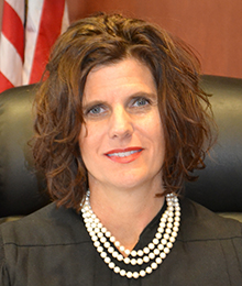 Honorable Sandra Upchurch Honorable Sandra Upchurch headshot, Caucasian woman with short brown hair and a pearl necklace is pictured in a large leather chair with an American flag behind her