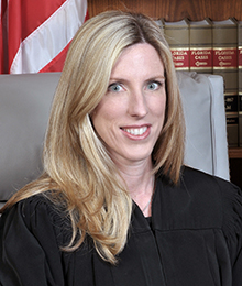 Honorable Stacy Ross Honorable Stacy Ross headshot, middle aged Caucasian female with blonde hair is seated on a large office chair with law books and an American flag in the background