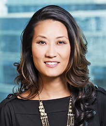 Katie Phang Katie Phang headshot, Asian businesswoman with long dark hair is wearing a black top with a statement necklace, pictured in front of a blue background