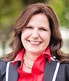 Paige Greenlee Paige Greenlee headshot, Caucasian female, middle aged with brown hair, wearing a black and white blazer with a red button down shirt and a pearl necklace